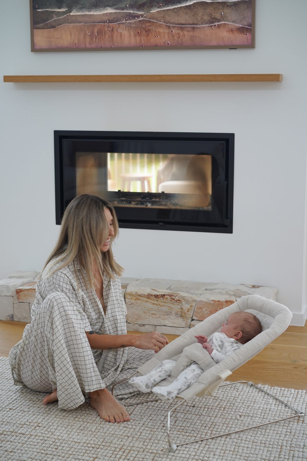 Mother kneeling beside baby in a bouncer seat near a fireplace