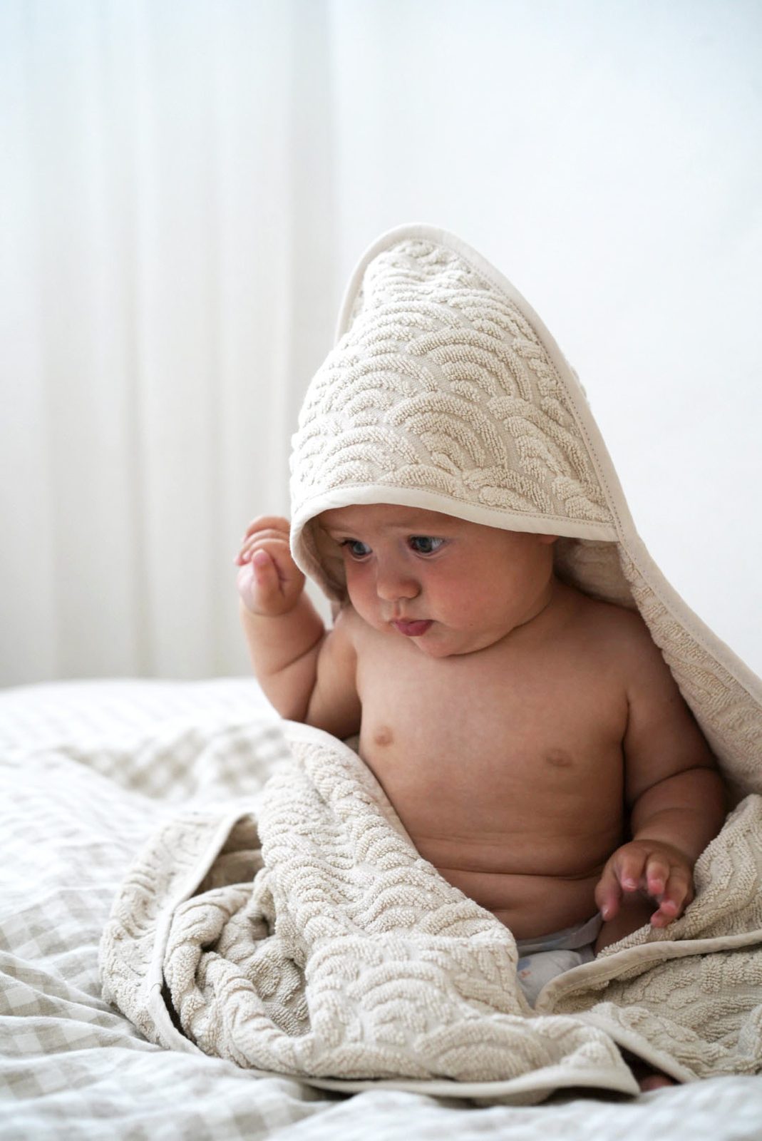 Baby wrapped in a textured cream hooded towel, sitting up and looking curiously to the side