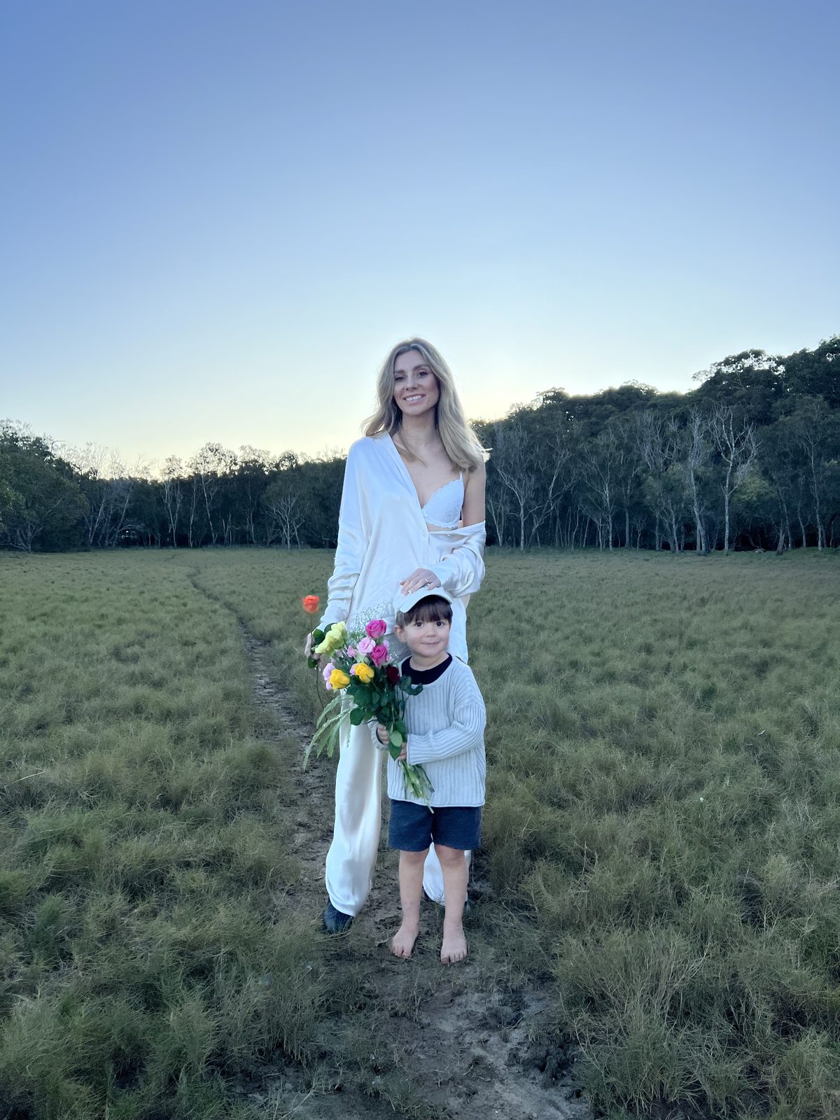 Mother holding her toddler on the beach at sunset while carrying a bouquet of flowers
