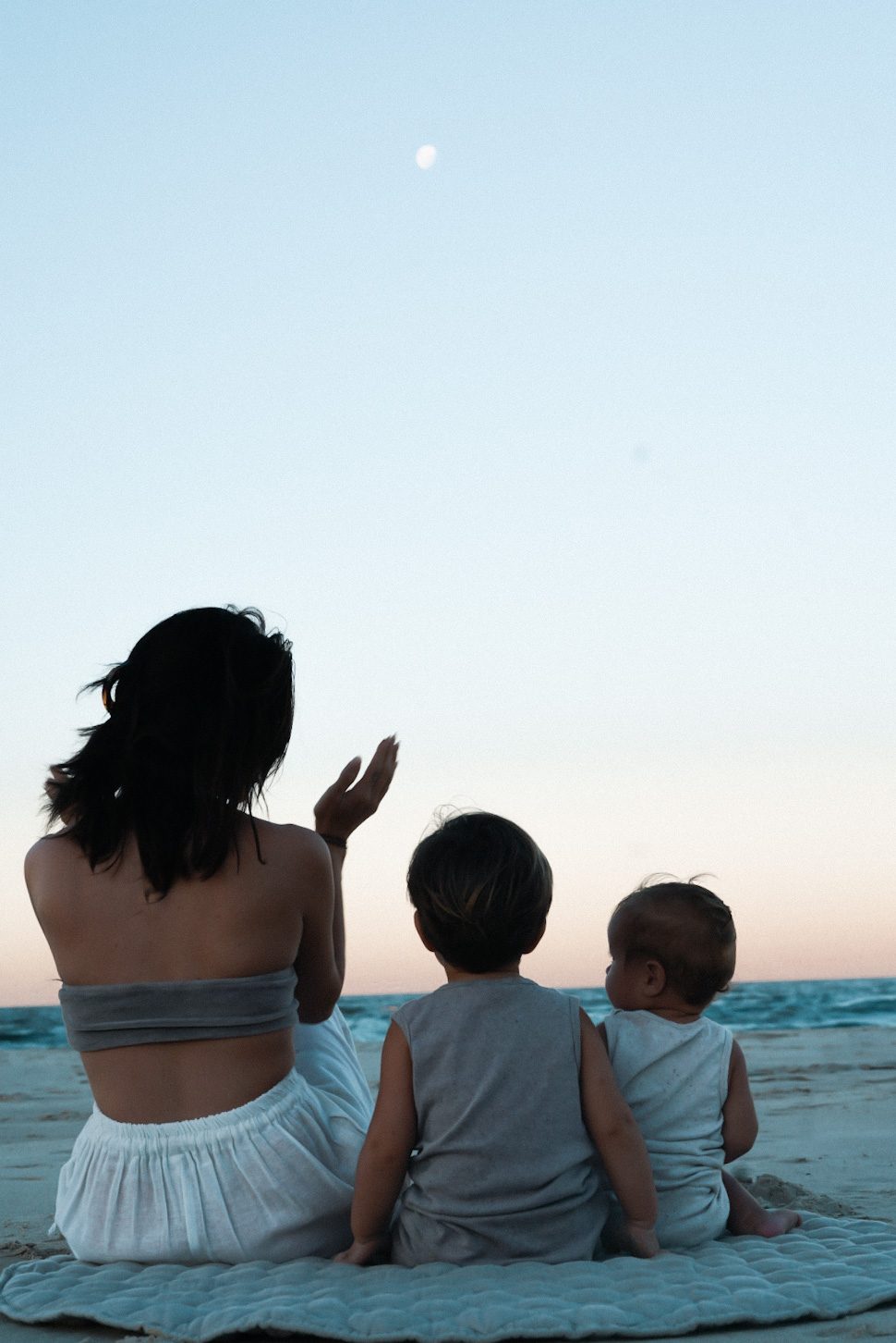 Mother and two young sons sitting on a beach blanket at dusk, watching the moon over the ocean