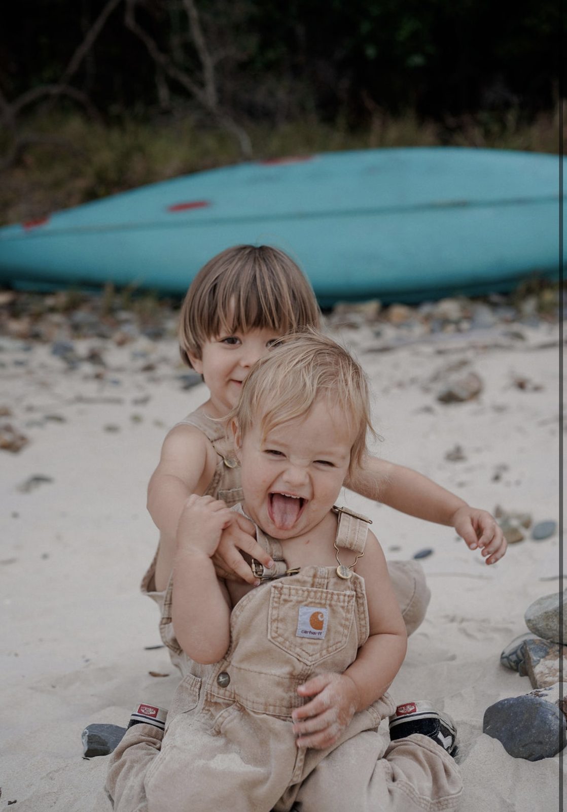 Two siblings in overalls laughing together on the beach with a blue kayak in the background