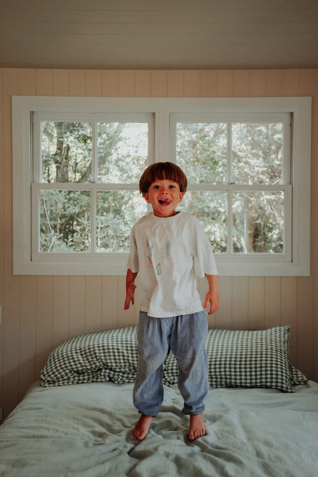 Boy sticking out his tongue while jumping on a bed in striped pyjama pants