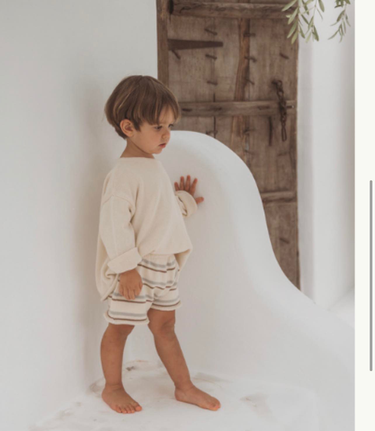 Toddler in a cream sweater and striped shorts standing beside a rustic wooden door
