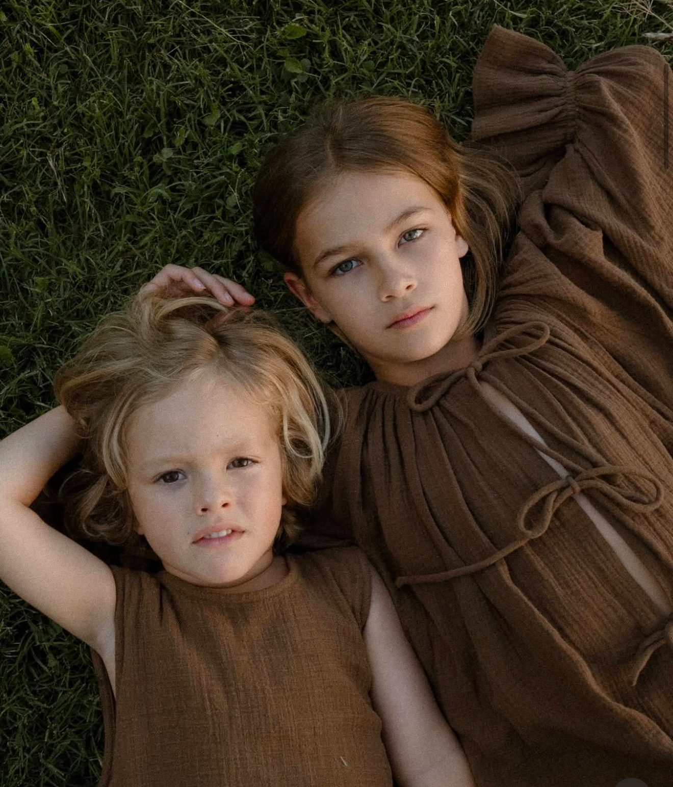 Two children lying in the grass wearing matching brown linen outfits