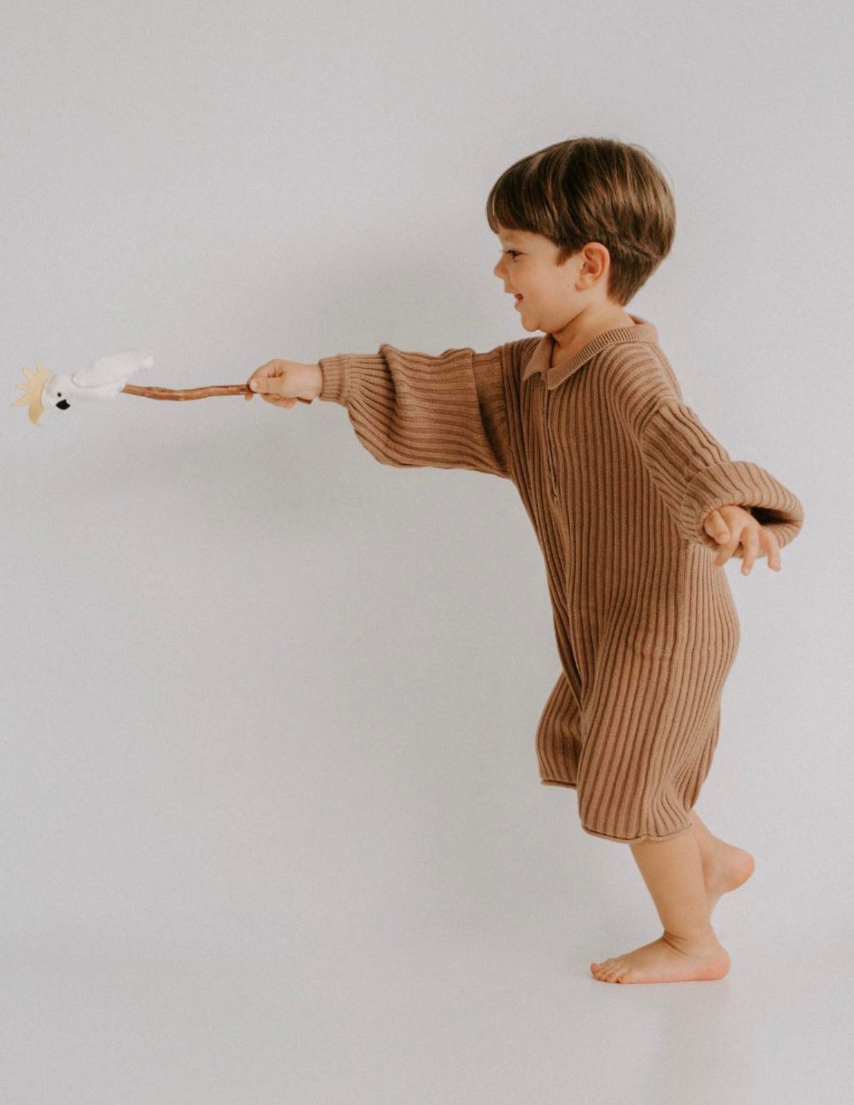 Toddler boy in a ribbed knit romper, playing with a felt cockatoo toy on a stick