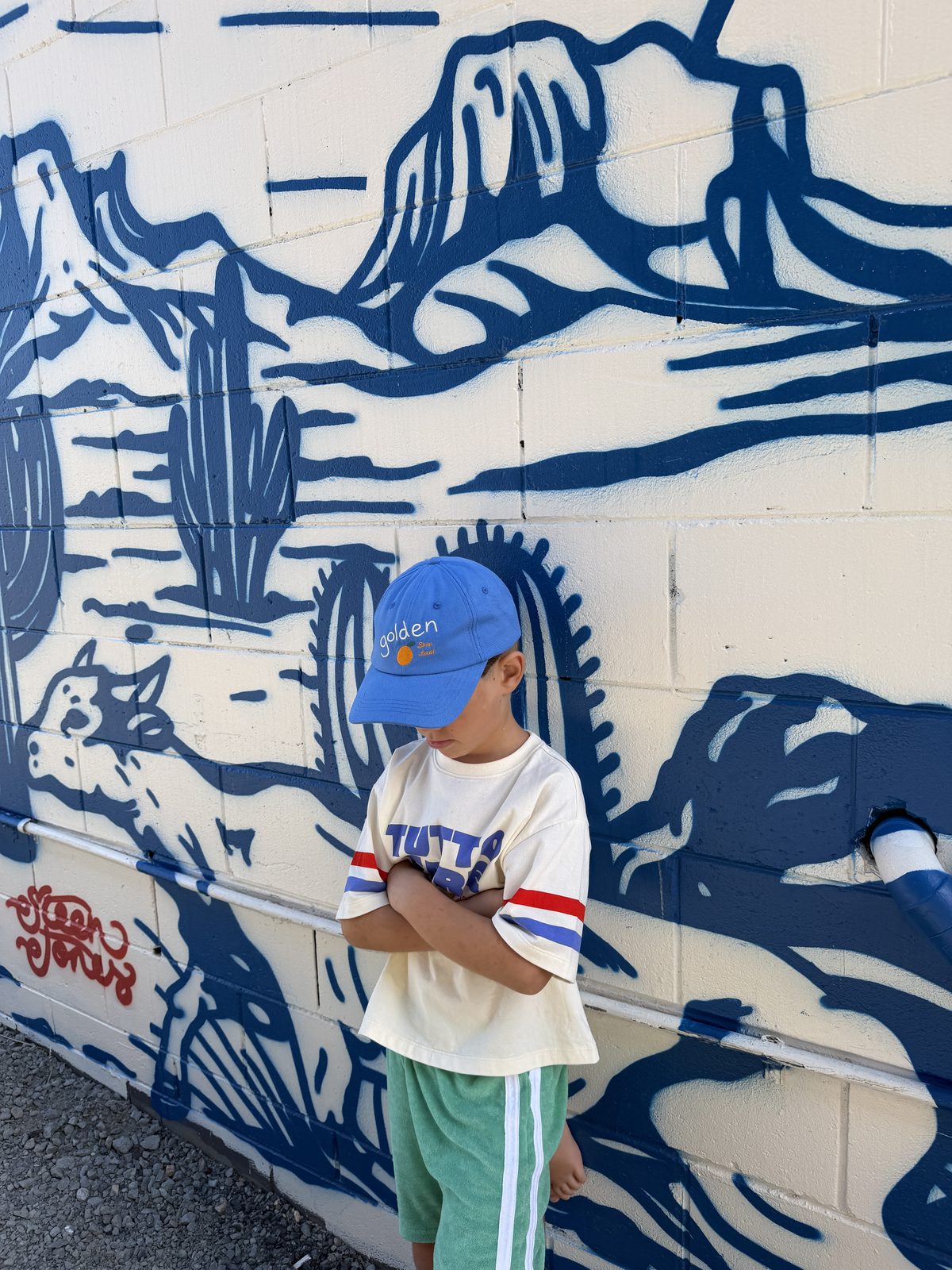 Toddler in a blue cap and cream striped tee standing in front of a blue mural
