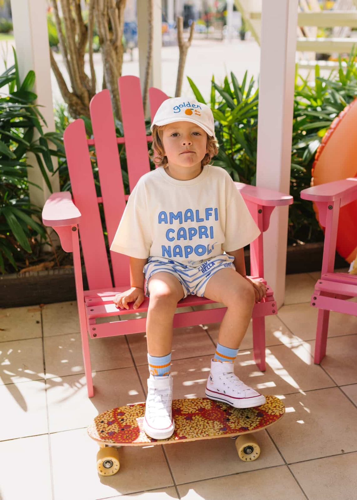 Young boy in an 'Amalfi Capri Napoli' graphic tee and cap standing on a skateboard