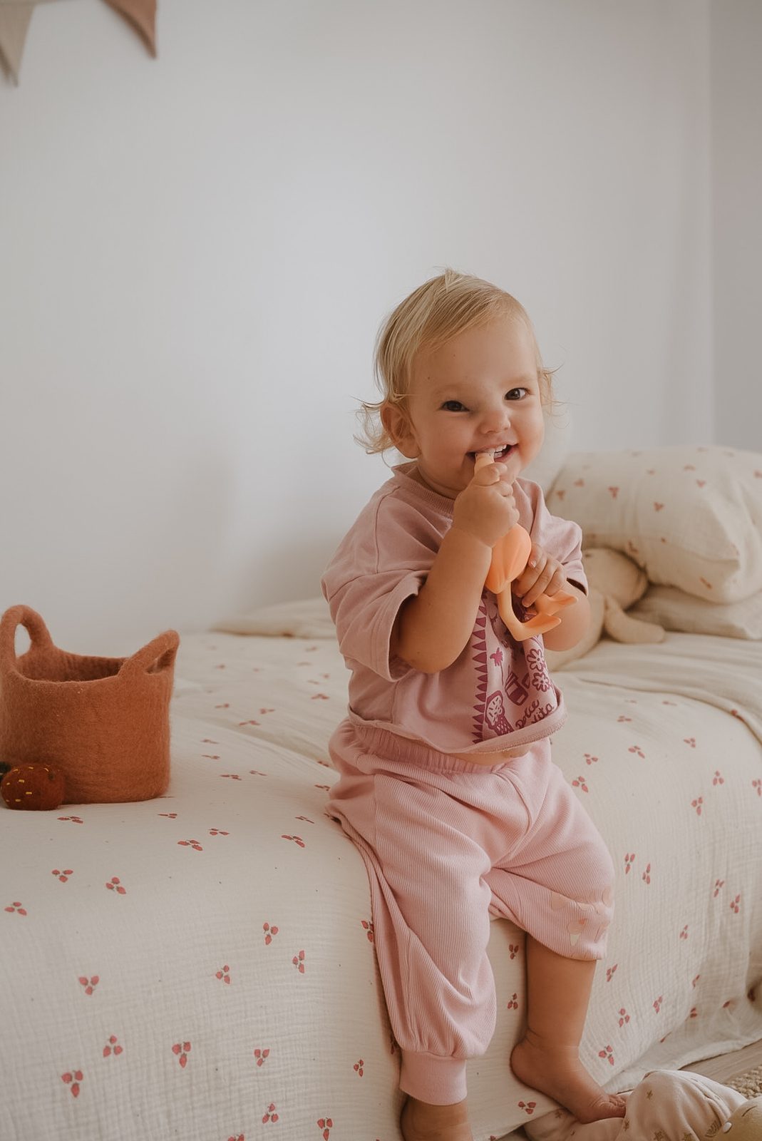 Toddler girl smiling while holding an orange teether toy, sitting on a bed