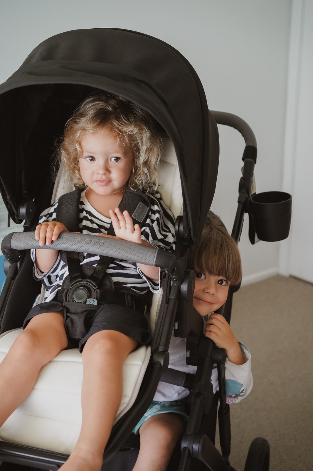 Two toddlers sitting together in a double stroller indoors