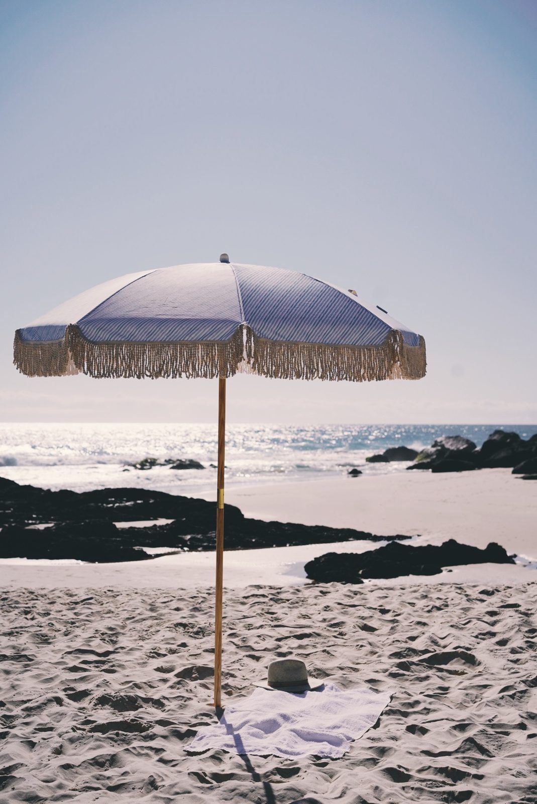 Fringed striped beach umbrella with a straw hat resting on a linen towel in the sand