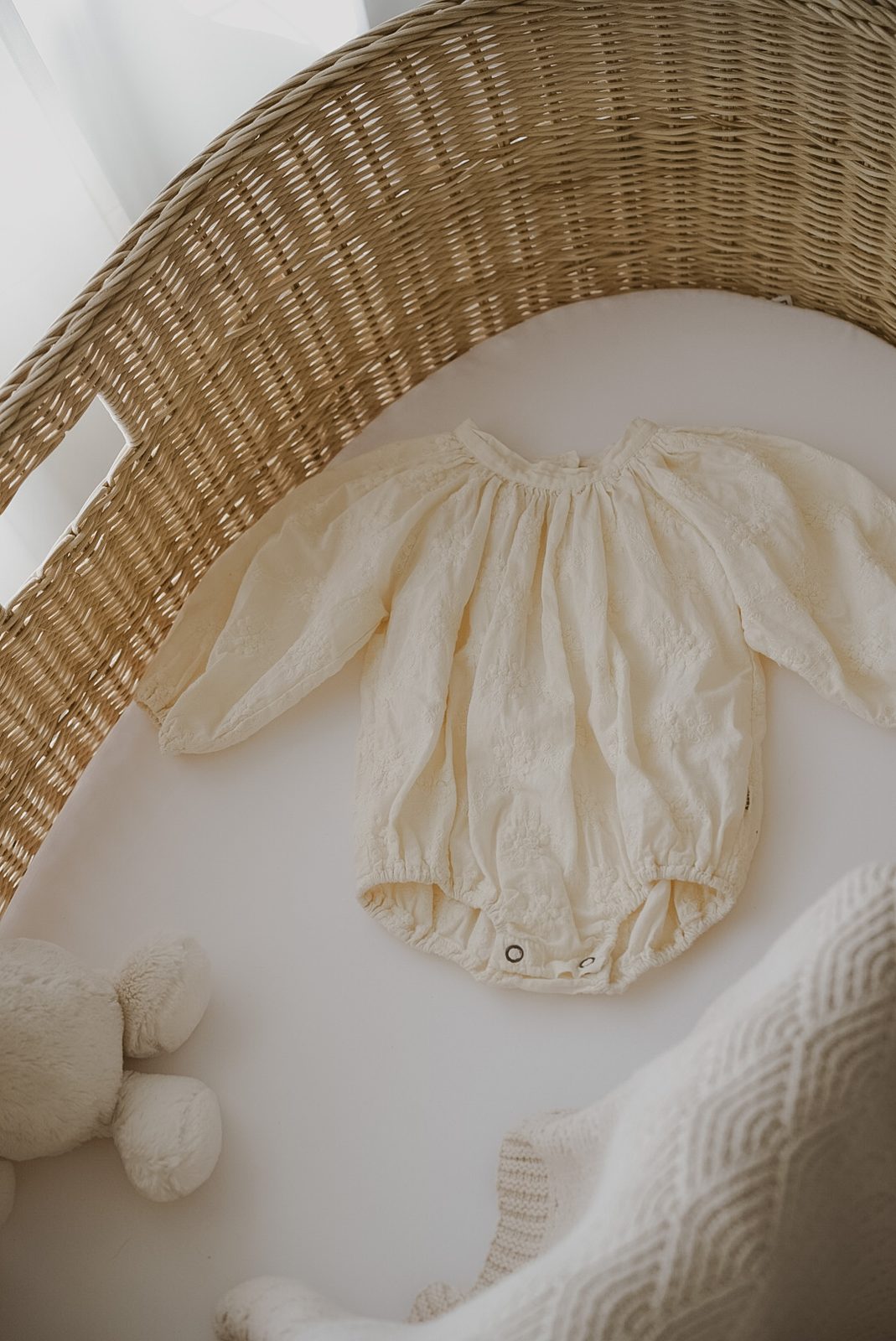 Cream embroidered baby romper laid out in a wicker bassinet with a plush toy